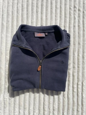 Mörkblå ziptröja från Morris - Mörkblå ziptröja från Morris med hög krage och hel dragkedja framtill. Tröjan har ribbade muddar och ett diskret broderat emblem på bröstet. Materialet är mjukt och bekvämt, perfekt för lager-på-lager och chill dagar.
