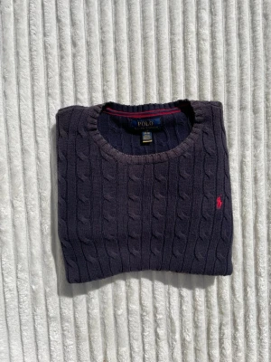 Mörkblå kabelstickad tröja Polo Ralph Lauren - Kabelstickad mörkblå tröja från Polo Ralph Lauren med rund halsringning och klassisk röd broderad logga på bröstet. Tröjan har ribbade muddar vid ärmslut och nederkant. Perfekt för dig som gillar tidlös och stilren design.