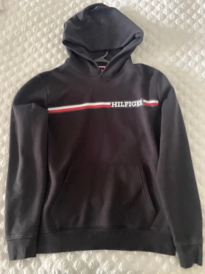 Svart hoodie från Tommy Hilfiger - Svart hoodie från Tommy Hilfiger med klassisk huva och magficka. På bröstet finns en vit, röd och blå rand samt Hilfiger-loggan broderad. Mjuk bomullskänsla och ribbade muddar vid ärmslut och nederkant. Perfekt för en avslappnad och stilren look.