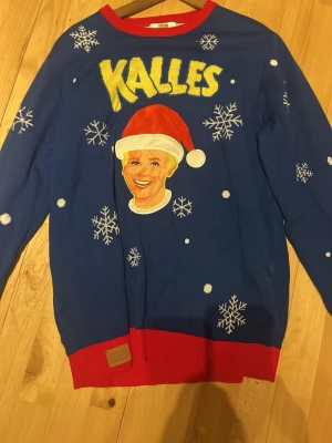 Blå jultröja Kalles x Svensk Husman - Rolig blå stickad jultröja från Kalles x Svensk Husman med snöflingor, röd tomteluva och gul text. Tröjan har röda muddar och halsringning samt ett broderat märke nertill. Perfekt för dig som gillar unika jultröjor!