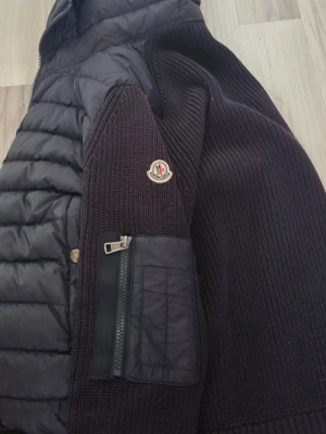 Mörkblå Moncler cardiganjacka - Snygg svart cardiganjacka från Moncler med quiltade partier och stickade ärmar. Jackan har dragkedja på ärmen, ribbstickade muddar och Moncler-logga på armen. Perfekt för dig som vill ha en trendig och bekväm jacka med exklusiv känsla.
