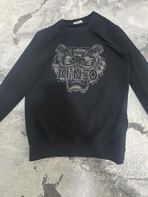 Svart sweatshirt från Kenzo - Svart sweatshirt från Kenzo med ikoniskt tigerhuvud och logga broderat på bröstet. Tröjan har rund halsringning, långa ärmar och ribbade muddar vid ärmslut och nederkant. Tillverkad i mjukt bomullsmaterial som är skönt mot huden.