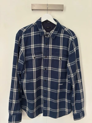 Blåvit rutig skjorta från Denham - Snygg blå och vit rutig skjorta från Denham med regular cut. Skjortan har långa ärmar, klassisk krage och knäppning framtill. Perfekt för dig som gillar en avslappnad men stilren look.
