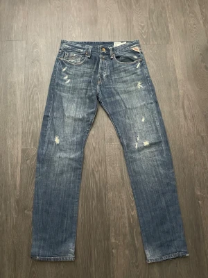Blåa replay jennon jeans - Säljer ett par blå replay jennon distressed jeans med raka ben och slitna detaljer fram och bak. Jeansen har klassisk femficksdesign och ljus tvätt med slitningar på lår och knän. Perfekta för en avslappnad och trendig look. Waist 31 length 34