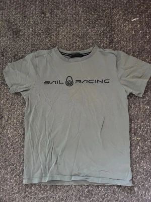 Grå Sail Racing t-shirt - Snygg grå/grön t-shirt från Sail Racing med svart logga och text framtill. Klassisk rund halsringning och korta ärmar. Tillverkad i mjuk bomull som känns skön mot huden. Perfekt för en avslappnad och sportig stil.
