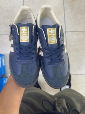 Adidas Samba mörkblå sneakers - Säljer ett par Adidas Samba sneakers i mörkblått med vita detaljer och klassiska tre ränder på sidan. Skorna har gummisula, mockaliknande material på tån och ovandel i textil och syntet. Guldiga detaljer på sidan och plös för extra stilpoäng.