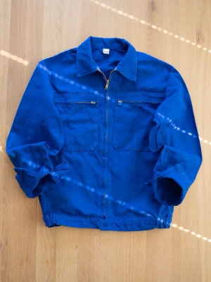 Vintage workwear jacka med dragkedja - Klassisk blå workwear jacka i bomull med två bröstfickor med dragkedja och hel dragkedja framtill. Jackan har en rak passform, nedfälld krage och långa ärmar. Perfekt för en avslappnad och trendig look.