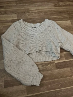 Beige cropped stickad tröja - Trendig beige stickad tröja med croppad passform och bred ribbad mudd vid halsringning, ärmslut och nederkant. Tröjan har ballongärmar och är gjord i ett mjukt, grovt stickat material. Perfekt för lager på lager och en avslappnad look.