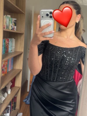 Svart långklänning med paljetter - Elegant svart långklänning med offshoulder-design och glittriga paljetter på överdelen. Klänningen har en åtsittande passform upptill och faller snyggt längs kroppen. Perfekt för bal eller festliga tillfällen.