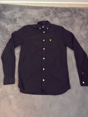 Svart skjorta från Lyle & Scott - Svart skjorta från Lyle & Scott med klassisk krage och vita knappar. På bröstfickan finns den ikoniska gula örnloggan broderad. Skjortan är långärmad och har en stilren, enkel design som passar till många olika outfits.