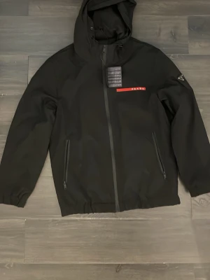 Svart vindjacka  - Snygg svart vindjacka med huva och dragkedja framtill. Jackan har två snedställda fickor med dragkedja och en röd logga på bröstet. Materialet är troligtvis polyester eller nylon, perfekt för en stilren och sportig look.