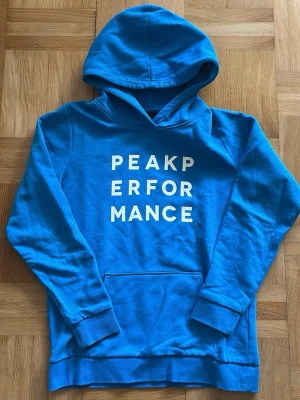 Blå Peak Performance hoodie - Snygg blå hoodie från Peak Performance med vit text framtill. Klassisk modell med huva och magficka. Perfekt för en avslappnad stil och enkel att matcha med jeans eller joggers. Mjuk bomullskänsla och ribbade muddar vid ärmslut och nederkant.