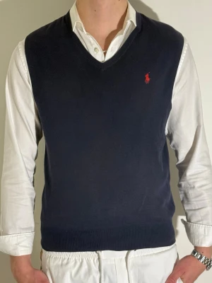 Mörkblå väst från Ralph Lauren - Klassisk mörkblå väst från Ralph Lauren med v-ringning och röd broderad logga på bröstet. Västen är i mjuk bomull och har ribbade kanter för en snygg passform. Perfekt att bära över skjorta för en stilren look.