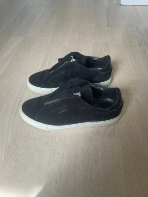Svarta Axel Arigato sneakers med dragkedja - Svarta sneakers från Axel Arigato i mocka med vit sula och cool dragkedja framtill istället för klassiska snören. Snygg minimalistisk design med diskret logga på sidan. Perfekta för dig som gillar stilrena och trendiga skor.