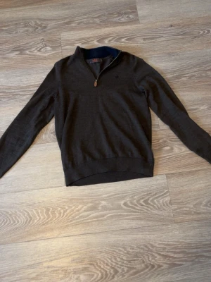 Morris tröja - Mörkbrun stickad tröja med halv dragkedja och liten mörkblå broderad detalj på bröstet. Tröjan har ribbade muddar vid ärmslut och nederkant samt en klassisk, stilren look. Perfekt för lager på lager och passar till jeans eller chinos.