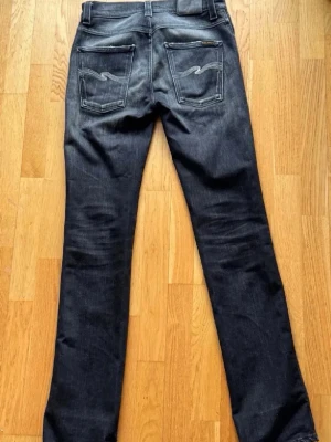 Mörkgråa Nudie Jeans - Snygga mörkgra jeans frän Nudie Jeans i modellen Black Lotus. Slim fit med klassisk femficksdesign som ger en modern, stilren silhuett. Naturliga slitningar ger varje par en unik vintagekansla, medan diskreta kontrastsömmar på bakfickorna forstarker den ikoniska Nudie-stilen