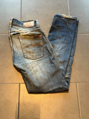 Blå slitna jeans från Nudie Jeans - Säljer ett par blå jeans från Nudie Jeans med tydliga slitningar och trasiga detaljer vid fickor och benslut. Jeansen har klassisk femficksmodell och smal passform. Perfekta för dig som gillar en avslappnad och trendig streetstil.