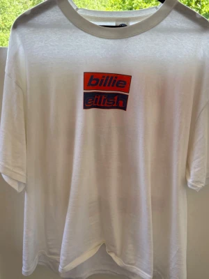 Vit Billie Eilish t-shirt med tryck - Billie Eilish t-shirt med tryck på både fram- och baksidan. Framsidan har logotypen i rött och blått, medan baksidan har ett stort grafiskt motiv och text. Den är aldrig använd! 