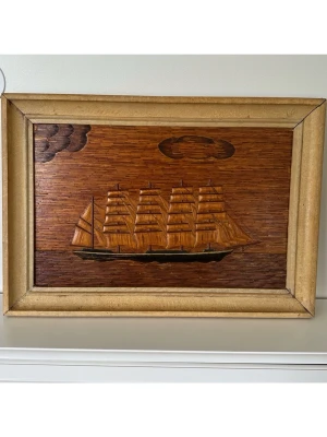 Trätavla med segelbåt, 66x46cm - Unik trätavla med motiv av en fyrmastad segelbåt, tillverkad i trä med inläggningar som ger en 3D-effekt. Tavlan har en bred, beige träram och motivet visar hav, moln och ett klassiskt skepp i bruna och gyllene toner. Mått 66x46cm. Vackert detaljerat hantverk i trä, unik i sitt slag. Perfekt som dekoration för dig som gillar marina teman. Fint skick!