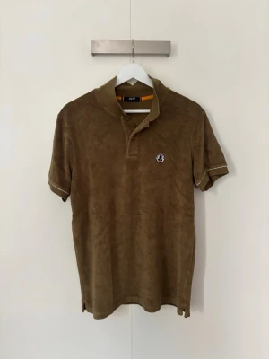 Brun polotröja från JOTT - Snygg brun polotröja från JOTT med korta ärmar och klassisk krage. Tröjan har en liten broderad logga på bröstet och är tillverkad i ett mjukt material som ger en avslappnad look. Perfekt till jeans eller shorts.