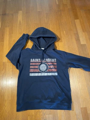 Mörkblå Saint Laurent hoodie - Mörkblå hoodie från Saint Laurent med tryck framtill i vitt och rött. Klassisk huva med dragsko och stor magficka. Trycket har texten 'SAINT LAURENT' och ett emblem med SL-logga. Perfekt för dig som gillar streetwear och vill ha en snygg, avslappnad stil.