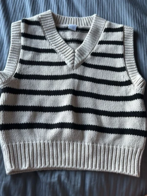 Randig stickad väst i offwhite och svart - Snygg stickad väst i offwhite med svarta horisontella ränder. Västen har v-ringning, bred ribbad kant nedtill och vid ärmöppningarna. Perfekt att bära över en skjorta eller t-shirt för en trendig lager-på-lager-look.