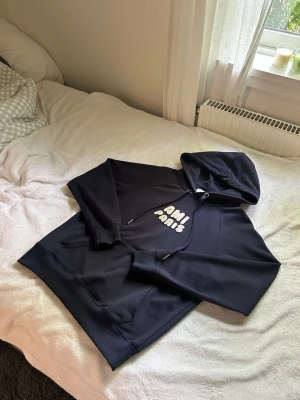Mörkblå hoodie från AMI Paris - Snygg mörkblå hoodie från AMI Paris med vit text på bröstet. Klassisk känguruficka framtill och justerbar huva med snören. Tillverkad i mjukt material som känns skönt mot huden. Perfekt för en avslappnad och trendig look.