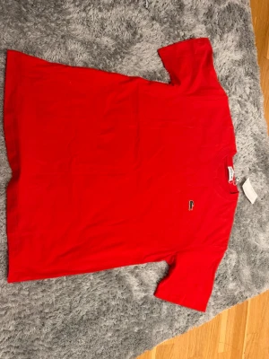 Röd Lacoste t-shirt i bomull - Klassisk röd t-shirt från Lacoste med den ikoniska krokodilloggan broderad på bröstet. T-shirten har korta ärmar och är tillverkad i mjuk bomull för en skön känsla. Perfekt till jeans eller shorts för en enkel och stilren look.