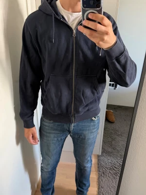 Mörkblå zip hoodie från Weekday - Mörkblå hoodie med dragkedja från Urban Outfitters. Klassisk modell med huva och dragsnören, två stora fickor framtill och ribbade muddar vid ärmar och nederkant. Tillverkad i mjuk bomullsblandning som är skön att ha på sig.