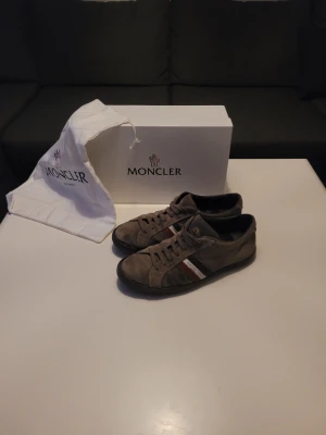 Moncler monaco skor - Snygga bruna sneakers från Moncler med klassiska ränder i rött, vitt och svart på sidan. Skorna har snörning och rund tå, samt en platt sula med Moncler-logga undertill. Utsidan är i mocka och insidan har vadderad skinnsula för extra komfort. Perfekt för dig som gillar stilrena detaljer. Skorna är lite limmade vid sulan