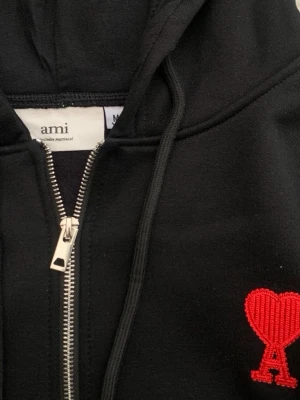 Svart hoodie med röd Ami-logo - Svart hoodie från Ami med dragkedja framtill och huva med snören. På bröstet finns Ami:s ikoniska röda hjärta med bokstaven A broderat. Hoodien har ribbade muddar och är tillverkad i mjukt material, perfekt för en chill och stilren look.