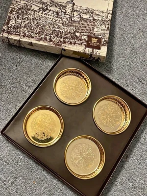 Scandia Guld glasunderlägg i presentask - Fyra runda glasunderlägg i äkta guldplätering från Scandia Present. Underläggen har ett detaljerat mönster och levereras i en snygg presentask med motiv av en stad. Perfekt för att ge en lyxig känsla till dukningen.