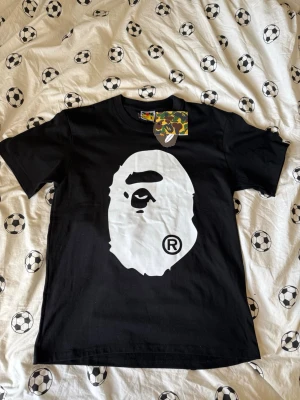 Svart BAPE t-shirt med stor logga - Säljer en svart t-shirt från BAPE med den ikoniska vita apansilhuetten tryckt stort på både fram- och baksidan. Klassisk passform och rund hals. Perfekt för dig som gillar streetwear och vill sticka ut med en tydlig logga.