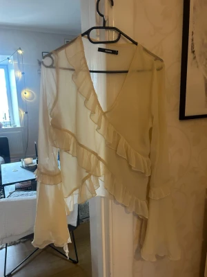 Beige omlottblus med volanger - Snygg beige omlottblus från bikbok i tunn, transparent chiffong. Blusen har långa ärmar med volangdetaljer och asymmetrisk skärning framtill. Perfekt för lager-på-lager och ger en romantisk vibe med sina volanger.