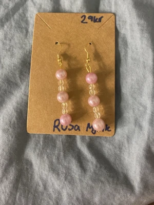 Rosa pärlörhängen med gulddetaljer - Säljer ett par handgjorda örhängen med rosa pärlor och små genomskinliga glaspärlor. Krokarna är guldfärgade och örhängena har en elegant, hängande design. Perfekta för dig som gillar söta accessoarer med en feminin touch. Alla örhängen e allergi fria