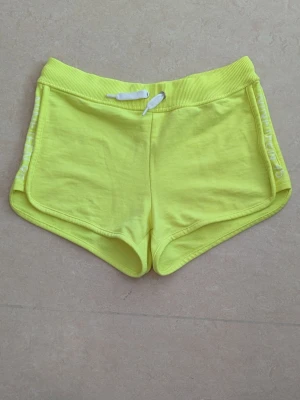 Gula shorts med snörning med tryckt text NINETEEN 95 - Snygga, sportiga shorts i neon-gul. De har vit snörning i midjan, ribbade kanter och tryckt text NINETEEN 95. Perfekta för sommaren eller träning, med mjukt material och avslappnad passform.,
