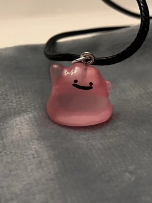 Självlysande Ditto halsband Pokémon smycke  - Så gulligt halsband med en berlock av en ljust rosa Ditto!Berlocken är ca 1,5 x 1,5 cm och trädd på svart tråd. Den är också självlysande och lyser i mörkret. Halsbandet är justerbart 40-45 cm.