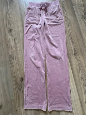 Rosa velourbyxor från Juicy Couture - Säljer ett par mjuka och sköna rosa velourbyxor från Juicy Couture. Byxorna har raka ben, bred resår i midjan med dragsko och två stora fickor fram. Klassisk Juicy Couture-logga broderad på ena fickan. Perfekta för en chill och trendig look.