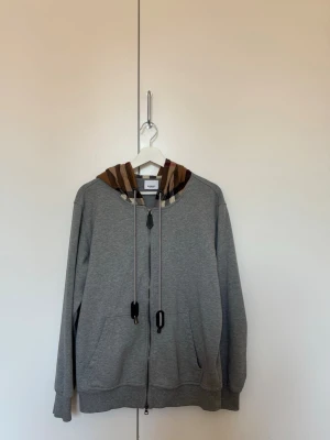 Burberry hoodie  - Säljer en grå zip hoodie från Burberry med klassisk beige, brun och röd rutig huva. Hoodien har dragsko i huvan, två fickor fram och ribbade muddar. Perfekt för dig som vill ha en stilren hoodie med en lyxig touch. Inga defekter nytt skick