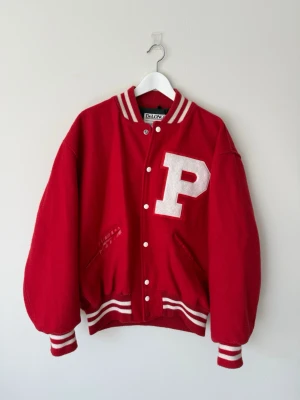 Amerikansk collagejacka - Äkta röd varsityjacka tillverkad i USA. Broderat P på bröstet och 'Balsley' på ryggen. Oversized passform där jag skulle säga att storlek är mer M-L. Yttre material är ull och nylon, inre är polyester.