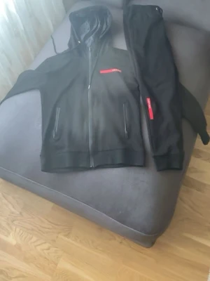 Svart tracksuit - Säljer ett par svarta mjukisbyxor från Prada med röd logotypdetalj på benet. Byxorna har en avslappnad passform och resår i midjan för extra komfort. Perfekta för en chill och stilren look.