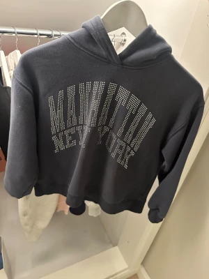 Svart hoodie med Manhattan-tryck - Cool svart hoodie med huva och texten 'MANHATTAN NEW YORK' i silvriga nitar på bröstet. Oversized passform och mjukt material som känns skönt mot huden. Perfekt för dig som gillar streetstyle och vill sticka ut lite extra.