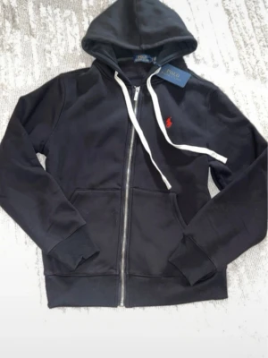 Svart hoodie från Polo Ralph Lauren - Svart hoodie med dragkedja från Polo Ralph Lauren. Klassisk design med huva, vita dragsnören och kängurufickor. Röd broderad logga på bröstet. Tillverkad i mjukt bomullsmaterial för en skön och avslappnad look.