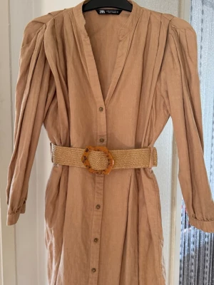 Beige linnéklänning med bälte från Zara - Säljer en beige långärmad klänning från Zara i linne med markerade axlar och snyggt bälte med stor spänne i midjan. Klänningen har knappar framtill och en avslappnad passform som ger en cool och trendig look.