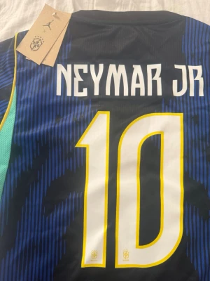 Brasilien Neymar JR matchtröja - Snygg Brasilien matchtröja med Neymar JR och nummer 10 på ryggen. Tröjan är svart med blå detaljer och gula inslag runt siffrorna. Tillverkad i lätt och ventilerande material, perfekt för fotboll eller träning. Officiell produkt med Brasil-logga och Jordan-märke.