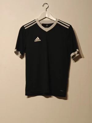 Adidas träningströja  - Snygg svart sporttröja från Adidas med vita detaljer på axlar, ärmslut och krage. Tillverkad i lätt och funktionellt Climalite-material som transporterar bort fukt. Perfekt för träning eller match. Klassisk Adidas-logga på bröstet.