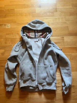 Grå hoodie från Burberry med dragkedja - Säljer en grå hoodie från Burberry med dragkedja och klassiskt rutigt foder i huvan. Hoodien har två stora fickor framtill, ribbade muddar och en skön, mjuk insida. Perfekt för dig som gillar stilrena och exklusiva plagg med snygga detaljer.