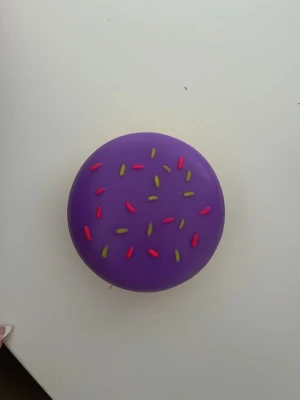 Nee Doh squishy i form av donut - Rolig squishy från Nee Doh som ser ut som en donut med lila ovansida, gul undersida och färgglada strössel. Tillverkad i mjukt gummiaktigt material, perfekt att klämma på när du behöver stressa ner eller bara vill ha något att pilla med.
