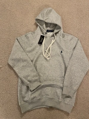 Grå hoodie från Polo Ralph Lauren - Snygg grå hoodie från Polo Ralph Lauren med klassisk huva och vita dragsnören. Tröjan har det ikoniska mörkblå polospelar-loggan broderad på bröstet och ribbade muddar vid ärmar och nederkant. Perfekt för en chill och stilren look.