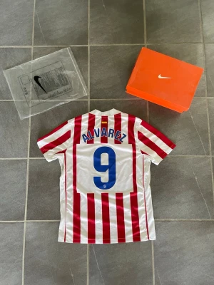 Atletico Madrid fotbollströja Nike - Säljer en Atletico Madrid fotbollströja från Nike med röda och vita vertikala ränder. Tröjan har nummer 9 och namnet Álvarez på ryggen samt Riyadh Air som sponsor på bröstet. Klassisk klubbsköld på vänster bröst och Nike-logga på höger.